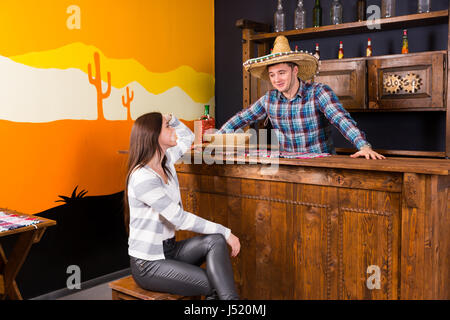 Eine junge Frau sitzt an der Bar neben der Barkeeper trägt Sombrero und ein kariertes Hemd in einer mexikanischen Bar entgegen Stockfoto