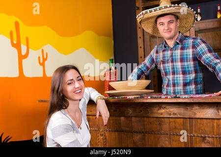 Glückliche Mädchen saß an der Bar neben der Barkeeper trägt Sombrero und ein kariertes Hemd in einer mexikanischen Bar entgegen Stockfoto