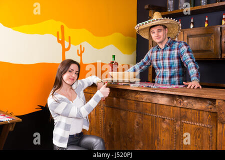 Junge Frau mit Daumen nach oben sitzen an der Bar neben der Barkeeper trägt Sombrero und ein kariertes Hemd in einer mexikanischen Bar counter Stockfoto