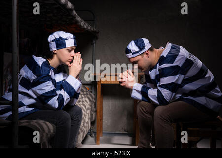 Junges Paar von Gefangenen Gefängnis tragen einheitliche sitzen in der Nähe von Nachttisch und beten in einer Gefängniszelle Stockfoto