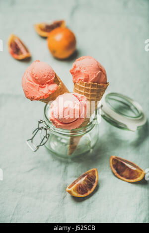 Erfrischende Sommer Blutorange Eis oder Sorbet Kugeln in süße Waffel Kegel im Glas serviert mit frischen Orangenscheiben, leichten grauen Tuch Hinterg Stockfoto