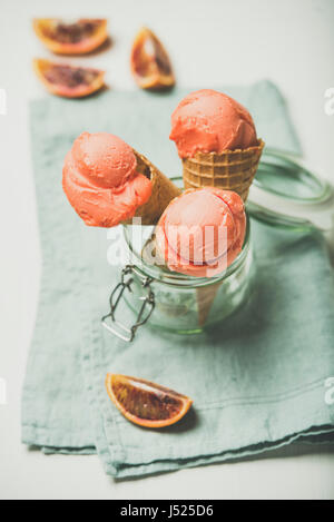 Erfrischende Sommer Blutorange Eis oder Sorbet Kugeln in süße Waffel Kegel im Glas serviert mit frischen Orangenscheibe, leichten grauen Tuch staatlich Stockfoto