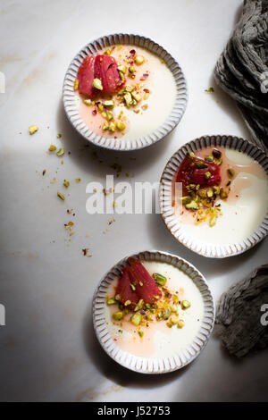 Klassische Vanille Pannacotta garniert mit gerösteten Rhabarber und gehackte Pistazien in einer keramischen Herb Pfanne auf einem Marmortisch in Draufsicht Stockfoto