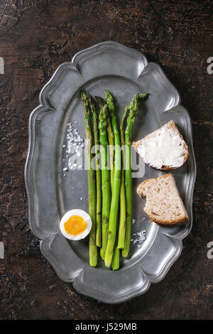 Gekochter grüner Spargel mit Hälfte gekochtes Ei und Schnittbrot mit Meersalz auf Vintage Metallplatte über braune Struktur Hintergrund serviert. Draufsicht, feine Stockfoto