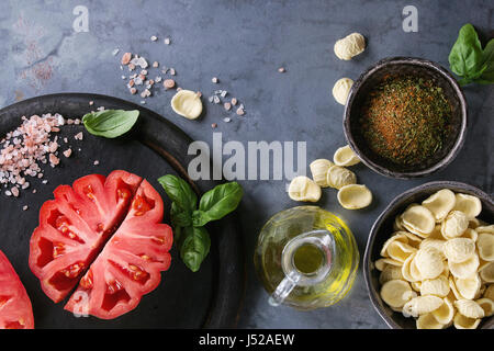 Hausgemachte rohe Teigwaren mit ganze und geschnittene Bio-Tomaten Coeur De Boeuf, Salz, Gewürze, Olivenöl und Basilikum über blau grau Metall Textur b Stockfoto