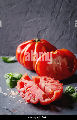 Ganze und geschnittene Bio-Tomaten Coeur De Boeuf. Beefsteak Tomaten mit rosa Salz und Basilikum auf blau grau Metall Textur Hintergrund. Nahaufnahme mit Raum Stockfoto