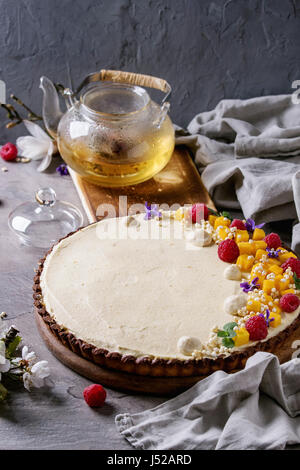 Hausgemachte Schokolade Tarte von Mango, Himbeeren, Minze dekoriert, aufgeblasen Reis und essbaren Blumen serviert mit Glas Teekanne und Textile Leinen über graue tex Stockfoto