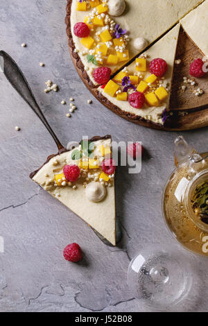 Hausgemachte geschnittene Schokolade Torte und Stück Tarte von Mango, Himbeeren, Minze dekoriert, aufgeblasen, Reis und essbaren Blumen serviert mit Glas Teekanne über g Stockfoto