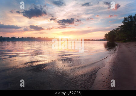 Farbenprächtigen Sonnenuntergang am Fluss. Sommerlandschaft mit Fluss, hellen Himmel mit Wolken und Sonne spiegelt sich im Wasser und Bäume am Strand. Natur Stockfoto