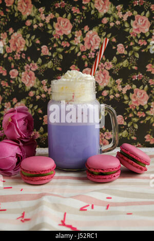 Lila Kaffee in Tasse stilisierte Einmachglas mit Macarons und Rosen und bunte Dekoration auf Hintergrund von floralen Muster. Heidelbeer-Milch-Shake. Einhorn Stockfoto