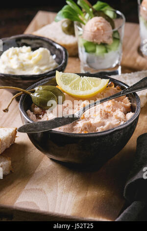 Schwarze Schale mit Lachspastete mit rotem Kaviar serviert mit Butter, in Scheiben geschnitten Brot, Kapern, Vintage Messer, Verrines und Kräutern auf hölzernen Portion Brett, Textil Stockfoto