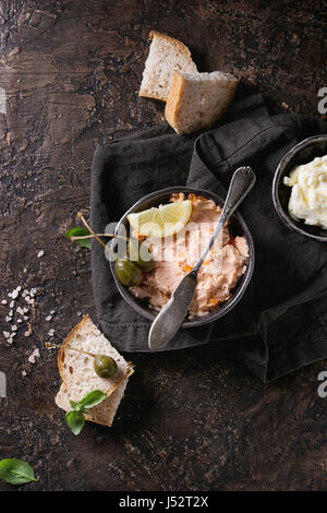 Schwarze Schale mit Lachspastete mit rotem Kaviar serviert mit Butter, in Scheiben geschnitten Brot, Kapern, Vintage Messer und Kräutern auf Textile Leinen Serviette über braune Textur Stockfoto
