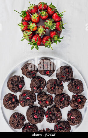 Schokoladen-Cookies mit Schokoladenstückchen und Erdbeeren auf einem weißen Teller und weißem Hintergrund begleitet von frischen Erdbeeren verpackt. Ansicht von oben mit Stockfoto