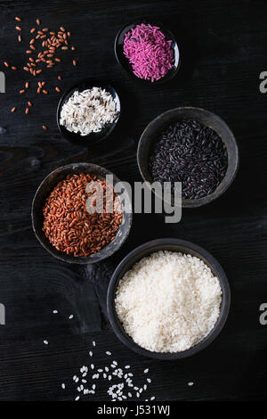 Verschiedene Auswahl an rohen ungekochten bunte Reis weiß, schwarz, braun, rosa, schwarz gebrannten Schalen über hölzerne Hintergrund. Draufsicht mit Raum Stockfoto