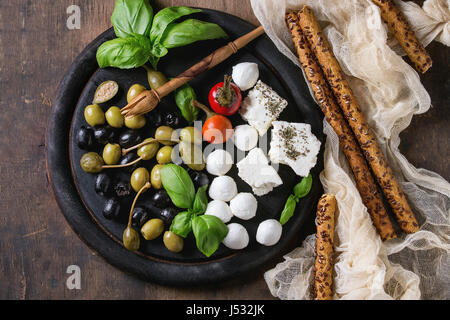 Mediterrane Vorspeise Antipasti-Board mit grünen und schwarzen Oliven, Feta-Käse, Mozzarella, Kapern, Pfeffer, Basilikum mit Grissini Brot über alte wo klebt Stockfoto