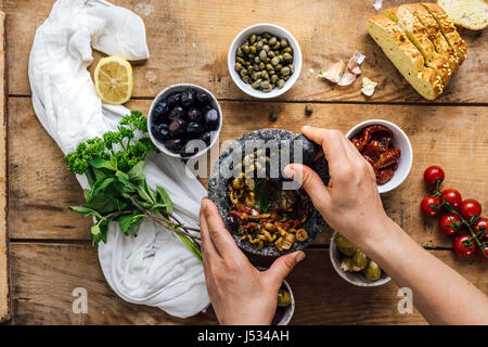 Eine Frau, die Oliven in einem Marmormörser Maischen zu Oliven Tapenade ist aus Draufsicht fotografiert.  Drei Arten von Oliven, Kapern, getrocknete Tomaten ihr Stockfoto