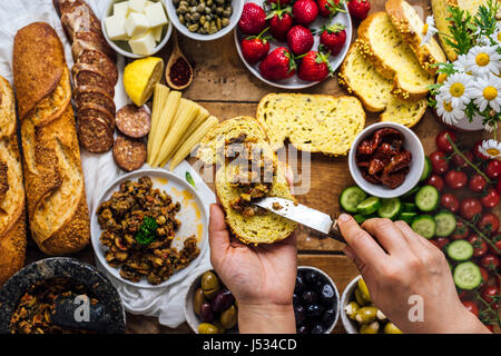 Eine Frau, die Oliven Tapenade auf einer Scheibe Maisbrot zu verbreiten. Französische Baguette, Wurst, Baby Hühneraugen, Käse, Strawberr, es, Blumen, Tomaten und Gurken Stockfoto