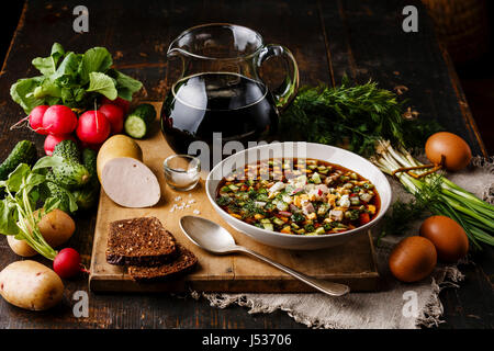 Kalte Suppe Okroschka mit Wurst, Gemüse und Kwas Portionsgröße in Schüssel und Rohstoffe auf Holztisch Stockfoto