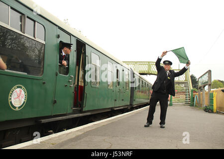 Swanage, UK - 12 Mai: Zug Wache winkt eine grüne Flagge an Corfe Castle Station vor der Dampf Züge Abfahrt Norden. Der l &amp; SWR 0-4-4 t Klasse M7 Nr. 30053 Dampflokomotive dauert 25 Minuten von Swanage, Norden verkehren. Gesamtansicht der Küste Stadt von Swanage in Dorset, England.  © David Mbiyu/Alamy Live-Nachrichten Stockfoto