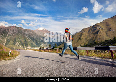 Tourist-Frau mit Rucksack und Regenbogen Hut liefen die Asphaltstraße in den Bergen in der Nähe von Big Almaty-See in Kasachstan Stockfoto