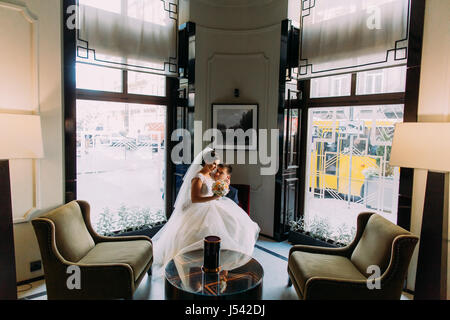 Charmante junge Braut sitzen auf Knien von ihrem schönen Bräutigam in einem luxuriösen Interieur mit glänzenden Fenstern als Hintergrund Stockfoto