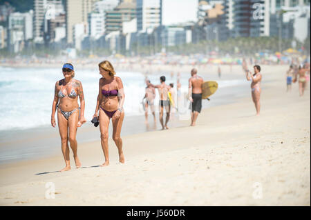 RIO DE JANEIRO - 9. Februar 2017: Strandbesucher in Bikinis zu Fuß am Ufer mit Teilen Surfer an einem hellen Sommernachmittag am Strand von Ipanema. Stockfoto