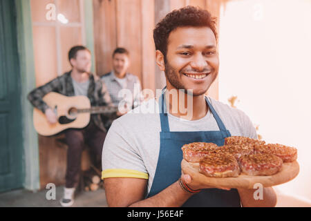 glücklicher Mann hält Burger auf Holzbrett, Freunde mit Gitarre sitzt hinter auf Veranda Stockfoto