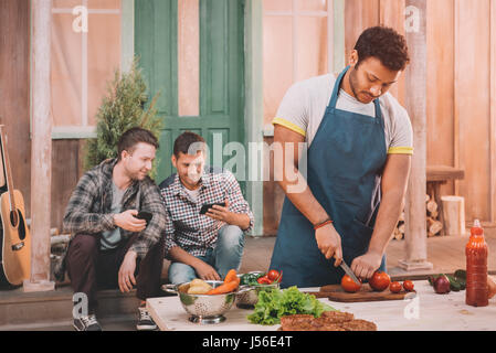 Mann, der Burger, während seine Freunde mit Smartphones sitzt hinter auf Veranda Stockfoto