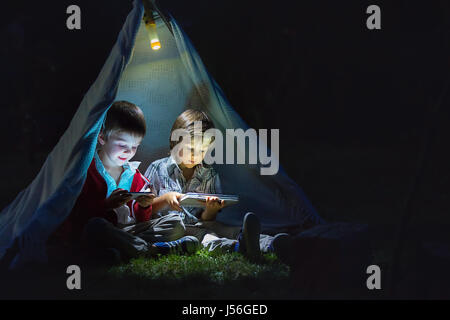 Niedliche kleine Brüder spielen auf Tablet und Telefon in der Nacht im Camp, im Zelt Stockfoto