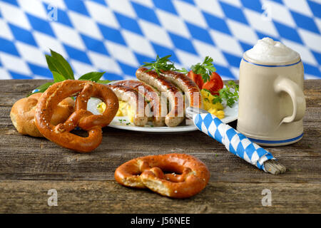 Gebratene bayerische Würstchen auf Kartoffelsalat serviert mit Brezeln und bayerisches Bier in einen Krug, im Hintergrund die weiß-blaue Flagge von Bayern Stockfoto