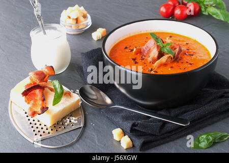 Dicke Tomatensuppe mit Basilikum und gebratenem Speck in einem schwarzen Keramikschale abstrakt grau unterlegt. Gesunde Ernährung-Konzept. Stockfoto