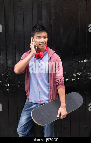 Porträt von einem asiatischen Mann mit seinem Smartphone und hält seinem Skateboard. Im Stadtstraße stehen im Freien. Stockfoto