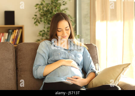 Stolz schwangeren Bauch sitzen auf einem Sofa im Wohnzimmer zu Hause mit einem warmen Licht durch die Fenster schauen Stockfoto