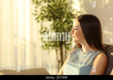 Seitenansicht einer Frau denken bei Sonnenuntergang sitzen auf einem Sofa im Wohnzimmer in einem Haus Interieur mit einem Fenster und ein warmes Licht im Hintergrund Stockfoto