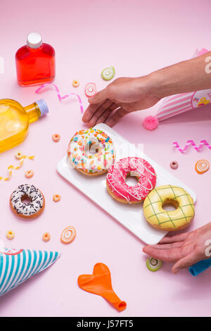Partei. Hand hält Teller mit bunten zuckerhaltige Runde glasierte Donuts und Flaschen Getränke auf rosa Hintergrund. Stockfoto
