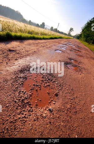 Lokalen roten Lehm Straße mit nassen Löchern umgeben von Feld und Wald. Schräg zu schießen. Polen, Woiwodschaft Świętokrzyskie. Stockfoto