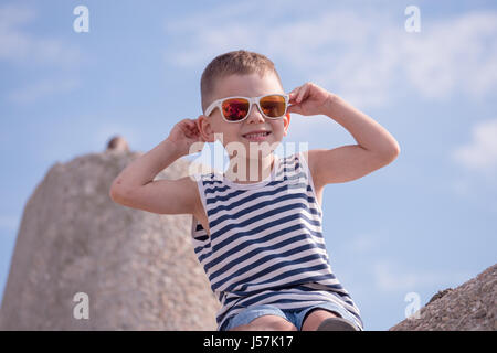 lächelnde Knabe mit Sonnenbrille und Streifen Matrosenhemd sitzen am Deich im Sommer Stockfoto