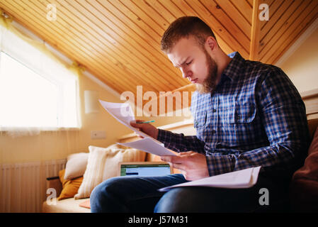 Junger Mann aus dem Hause sitzen auf einem Sofa hält Dokumente während der Arbeit an einem Laptop-Computer zu studieren. Stockfoto