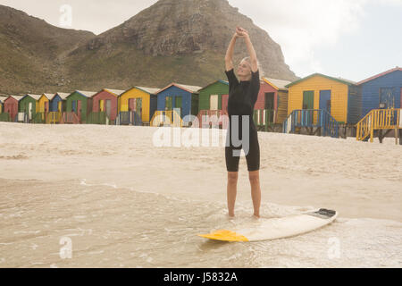 Frau ausdehnen, während sie von Surfbrett stehend gegen Hütten Stockfoto