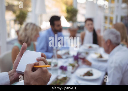 Nahaufnahme der Kellner schreiben Bestellung auf Notizblock während Freunde Essen im Hintergrund Stockfoto