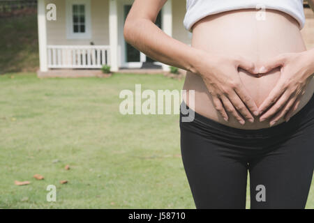 junge asiatische schwangere Frau die Form Herzschild auf ihrem Bauch vor ihrem Haus.  Schwangerschaft, Mutterschaft Bauch Pflegekonzept Stockfoto