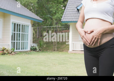 junge asiatische schwangere Frau die Form Herzschild auf ihrem Bauch vor ihrem Haus.  Schwangerschaft, Mutterschaft Bauch Pflegekonzept Stockfoto