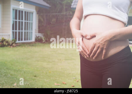 junge asiatische schwangere Frau die Form Herzschild auf ihrem Bauch vor ihrem Haus.  Schwangerschaft, Mutterschaft Bauch Pflegekonzept Stockfoto