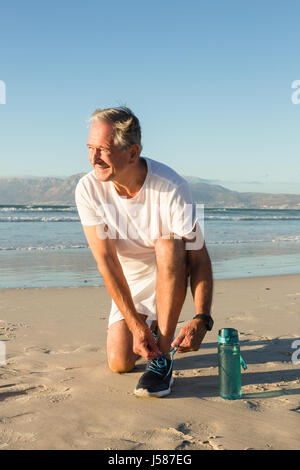 Lächelnder senior Mann binden Schnürsenkel am Strand gegen klaren Himmel Stockfoto