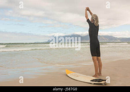 Ansicht der Rückseite Frau stretching während von Surfboard gegen bewölkter Himmel stehen Stockfoto