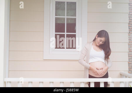 junge asiatische schwangere Frau die Form Herzschild auf ihrem Bauch vor ihrem Haus.  Schwangerschaft, Mutterschaft Bauch Pflegekonzept Stockfoto