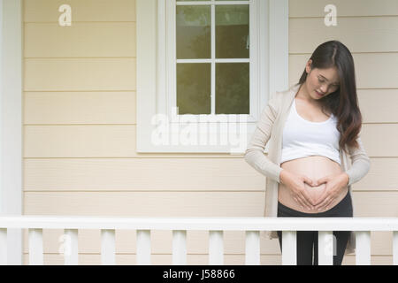 junge asiatische schwangere Frau die Form Herzschild auf ihrem Bauch vor ihrem Haus.  Schwangerschaft, Mutterschaft Bauch Pflegekonzept Stockfoto