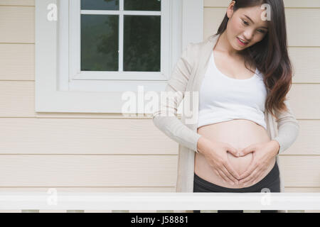 junge asiatische schwangere Frau die Form Herzschild auf ihrem Bauch vor ihrem Haus.  Schwangerschaft, Mutterschaft Bauch Pflegekonzept Stockfoto