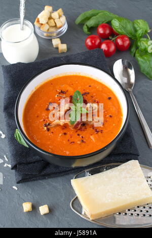 Dicke Tomatensuppe mit Basilikum und gebratenem Speck in einem schwarzen Keramikschale abstrakt grau unterlegt. Gesunde Ernährung-Konzept. Stockfoto