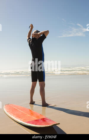 Rückansicht des Menschen tragen Neoprenanzug stehend von Surfbrett am Strand Stockfoto